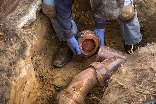 Man removing sections of old clogged clay ceramic sewer pipe in trench in the ground.